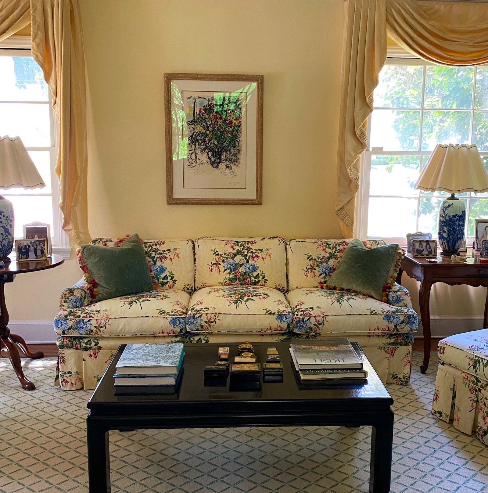 A bright, classic living room, featuring a custom-upholstered sofa, pleated lampshades, tassled velvet pillows, and pooled butter-yellow drapes, all inspired by California sunshine, and the fresh flowers of the treasured Marc Chagall framed artwork that anchors the room.